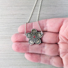 Load image into Gallery viewer, Silver flower-shaped necklace with a red gemstone held between fingers on a light gray background
