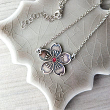 Load image into Gallery viewer, Silver floral pendant with red gemstone necklace on a textured ceramic stand
