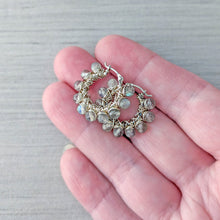 Load image into Gallery viewer, A pair of small hoop earrings with faceted gray gemstone beads wrapped in silver wire, held on a person's fingers against a light gray wood background.
