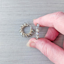 Load image into Gallery viewer, A close-up of a hand holding a silver hoop earring with light gray faceted stones with flashes of blue, next to another matching hoop earring lying on a gray wood-textured surface.
