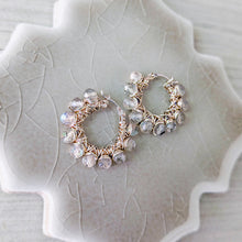 Load image into Gallery viewer, A pair of handmade hoop earrings with silver wire wrapping and grayish beige faceted stones with iridescent blue flashes, displayed on a textured ceramic leaf-shaped plate.
