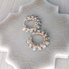 Load image into Gallery viewer, A pair of handmade hoop earrings with silver wire wrapping and grayish beige faceted stones with iridescent blue flashes, displayed on a textured ceramic leaf-shaped plate.
