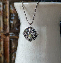 Load image into Gallery viewer, A silver spider with a smooth opal stone set into the abdomen hanging from a beaded silver chain emerging from a crackle glaze white vase with a blurred background
