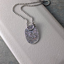 Load image into Gallery viewer, The reverse side of the pendant with a floral paisley pattern laying on a crackle glazed beige tile
