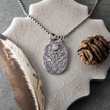 Load image into Gallery viewer, A silver necklace with an intricately designed pendant depicting a flying bat with a pale gray stone laying on a ceramic tile with a feather and a seed pod

