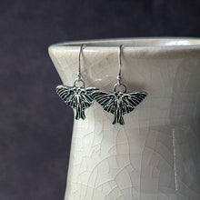 Load image into Gallery viewer, Silver Luna moth earrings on a textured white surface with a dark background
