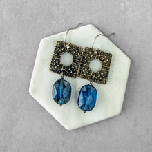 Load image into Gallery viewer, A pair of handmade dangle earrings displayed on a white hexagonal stand against a textured gray background. The earrings feature square, oxidized bronze-toned metal plates with a cutout center and dangling faceted blue gemstone beads.
