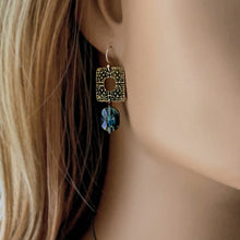Load image into Gallery viewer, A close-up of a woman's ear wearing a dangling earring with an ornate square gold-toned filigree charm and a faceted blue gemstone bead.
