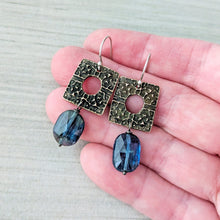 Load image into Gallery viewer, A pair of dangling earrings displayed on a person's hand. Each earring has an oxidized bronze square pendant with a circular cutout and a faceted dark blue oval gemstone dangling below.
