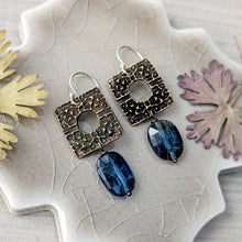 Load image into Gallery viewer, A pair of handmade dangle earrings displayed on a textured ceramic leaf-shaped plate with dried botanical leaves. The earrings feature textured antique bronze square charms with a central cutout, silver-tone ear wires, and a dangling faceted blue gemstone bead.
