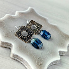 Load image into Gallery viewer, A pair of dangling earrings displayed on a textured ceramic leaf-shaped tray. The earrings feature square, oxidized bronze-toned metal plates with circular cutouts and oval blue faceted gemstones.
