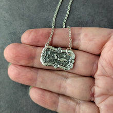 Load image into Gallery viewer, Silver necklace with a decorative pendant held in a hand against a gray background

