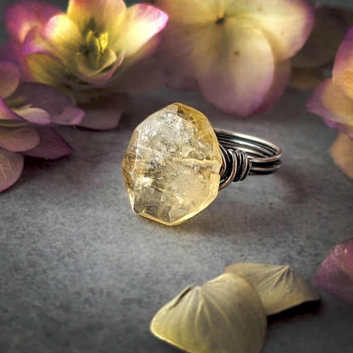Ring with a large, faceted yellow gemstone on a gray surface with flowers in the background
