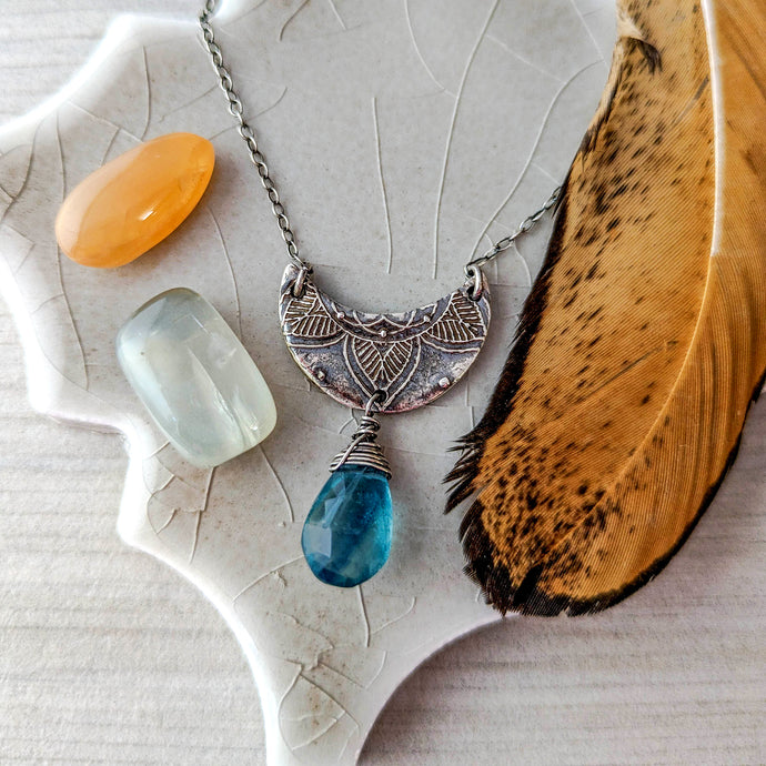 Silver crescent moon pendant with blue teardrop gemstone, orange and grey stones, and feather on textured surface