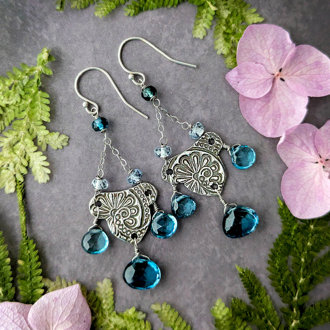 Silver earrings with London blue topaz briolettes on a gray surface with green leaves and pink flowers.
