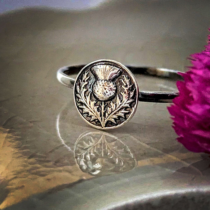 Silver ring with intricate thistle design on a reflective surface with a pink flower.