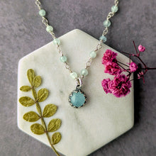 Load image into Gallery viewer, Necklace with a blue pendant on a hexagonal stone surface with dried leaves and pink flowers.
