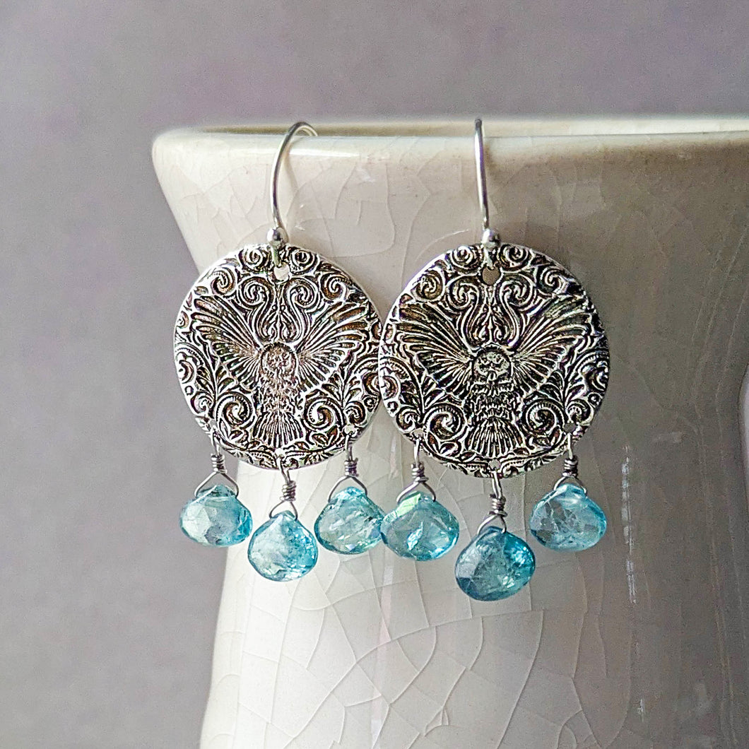 Silver earrings with an embossed owl design and blue teardrop gemstone beads on a white ceramic stand.