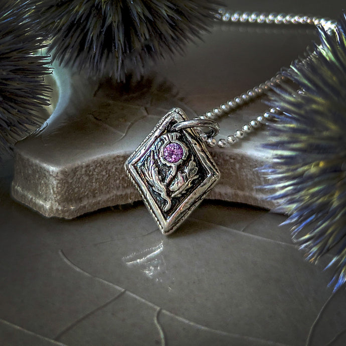 Fine silver thistle pendant with a pink sapphire on a ceramic surface with globe thistles in the foreground and background