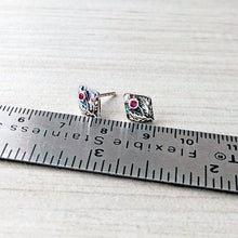 Load image into Gallery viewer, Silver diamond shaped stud earrings with dark pink gemstones on a ruler for scale on a white background
