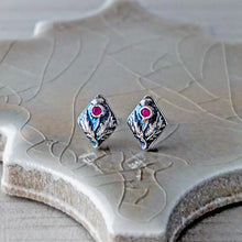 Load image into Gallery viewer, A pair of silver-toned diamond-shaped stud earrings with  floral design and a small pink gemstone, displayed on a textured ceramic tile.
