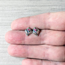 Load image into Gallery viewer, A pair of small diamond shaped silver stud earrings with pink gemstones in a floral design, shown resting on a person's fingers against a light gray textured background.
