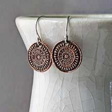 Load image into Gallery viewer, A pair of round copper-toned drop earrings with intricate circular medallion patterns, hanging from silver fishhook wires, displayed against a white ceramic vase.
