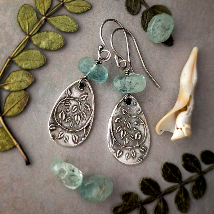 Silver earrings with blue green aquamarine gemstones on a textured surface with leaves and a shell.