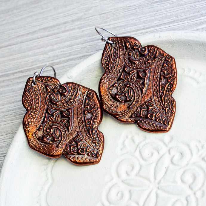 Ornate floral embossed copper earrings with an arabesque shape on a white background.
